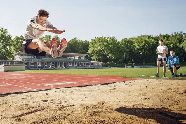 Weitspringer in der Luft beim Absprung über eine Sandgrube, daneben zwei Beobachterinnen.