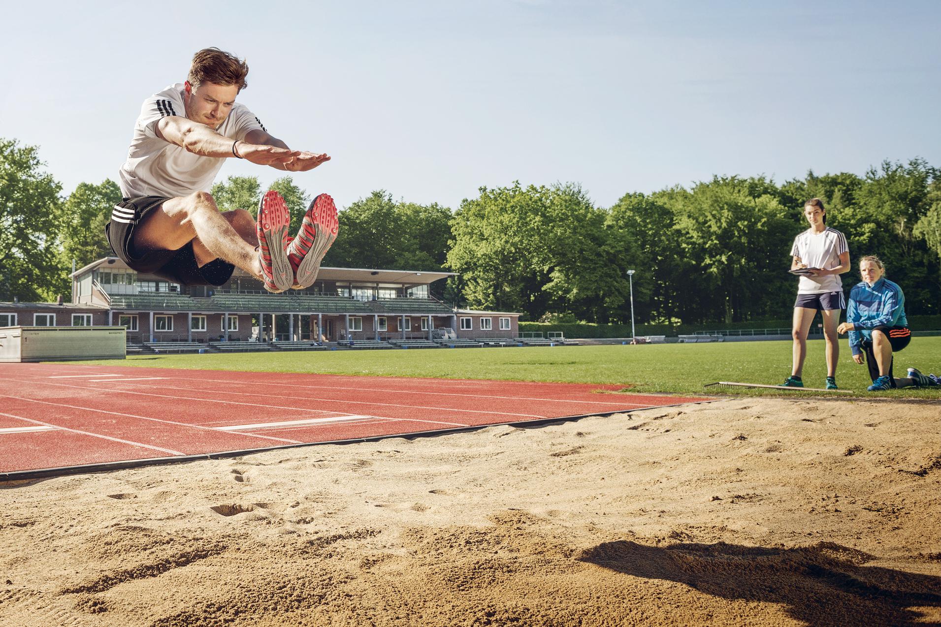 Weitspringer in der Luft beim Absprung über eine Sandgrube, daneben zwei Beobachterinnen.
