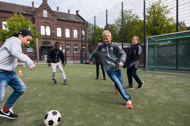 Fünf Kinder spielen Fußball auf einem Kunstrasenplatz, während ein Jugendgebäude im Hintergrund zu sehen ist.