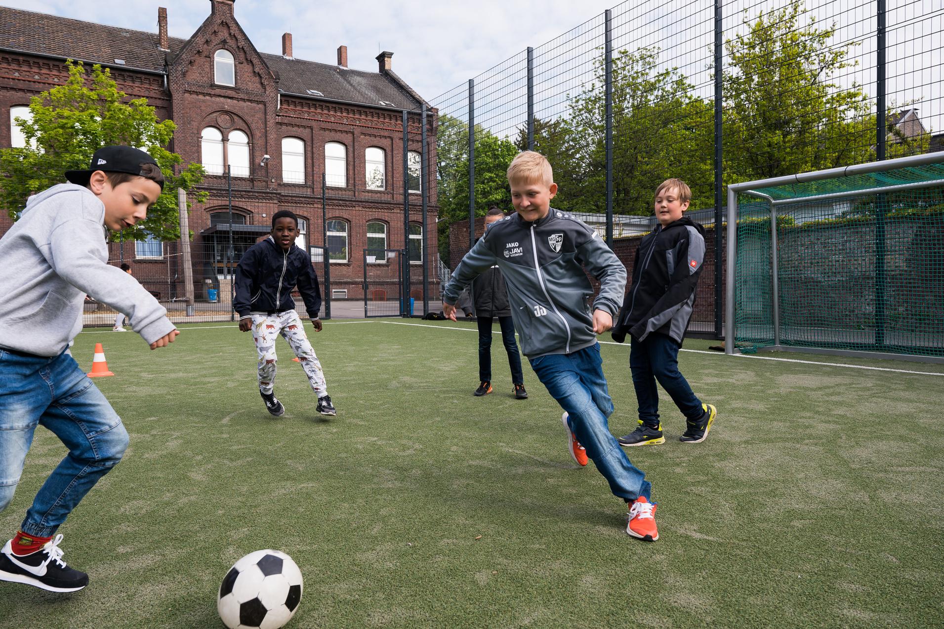 Fünf Kinder spielen Fußball auf einem Kunstrasenplatz, während ein Jugendgebäude im Hintergrund zu sehen ist.