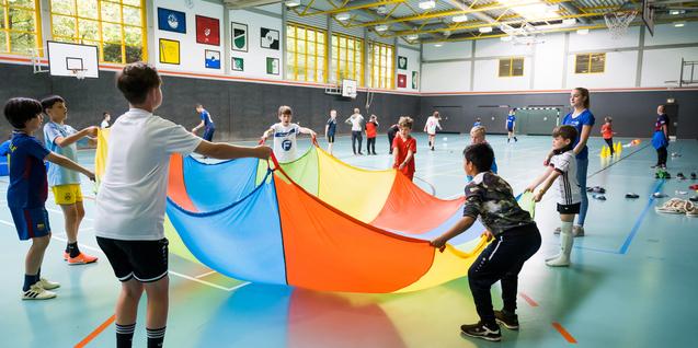 Gruppe von Kindern in einer Sporthalle, die einen bunten Parachute in die Höhe halten und gemeinsam spielen.