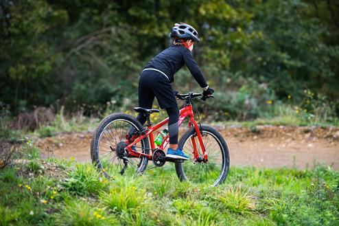 Junge auf einem roten Mountainbike fährt auf einem schmalen, unebenen Weg durch eine grüne Landschaft.