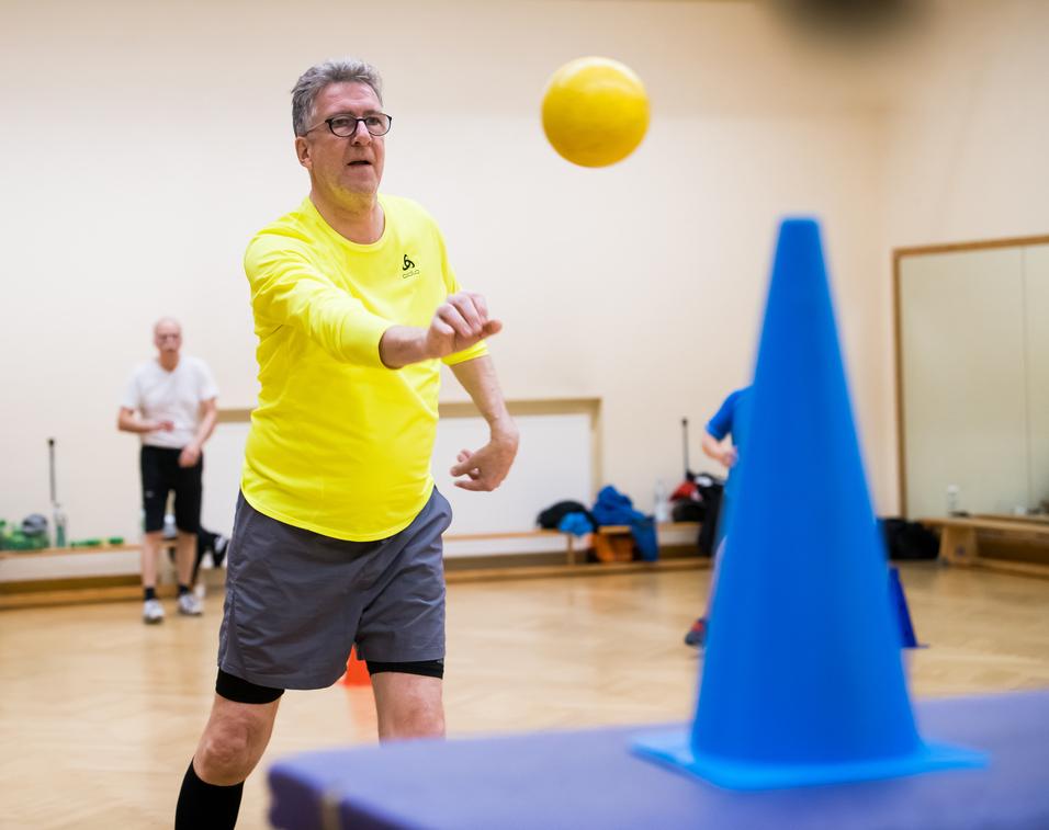 Ein Mann in gelber Sportkleidung wirft einen Ball auf einen blauen Kegel in einem Übungsraum. Hintergrund unscharf.