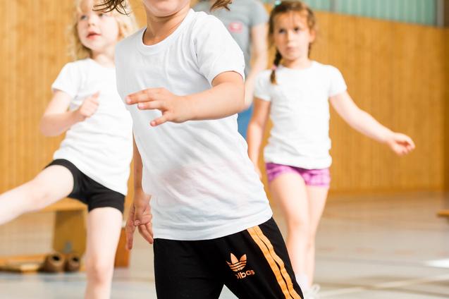 Vier Kinder in Sportkleidung bewegen sich in einer Turnhalle, ein Mädchen mit Zöpfen lächelt in die Kamera.