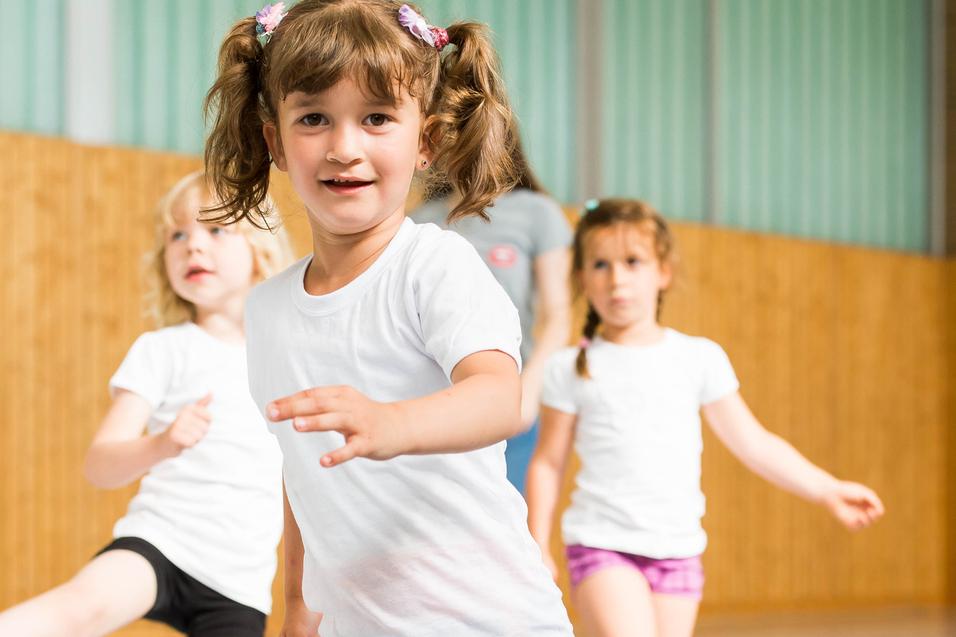 Vier Kinder in Sportkleidung bewegen sich in einer Turnhalle, ein Mädchen mit Zöpfen lächelt in die Kamera.