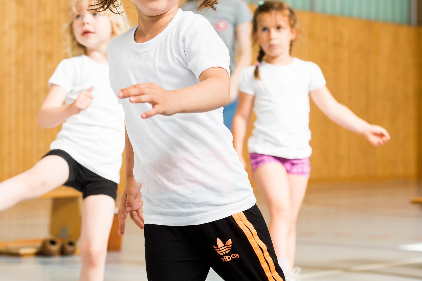 Vier Kinder in Sportkleidung bewegen sich in einer Turnhalle, ein Mädchen mit Zöpfen lächelt in die Kamera.