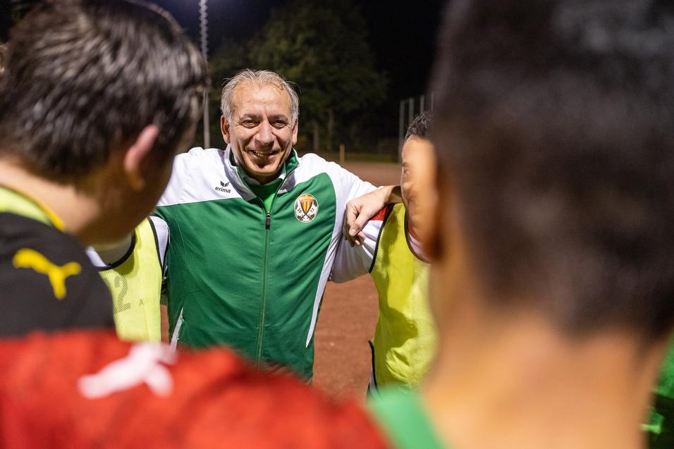 Trainer umringt von Spielern in sportlichen Trikots während einer Besprechung auf dem Fußballplatz bei Nacht.